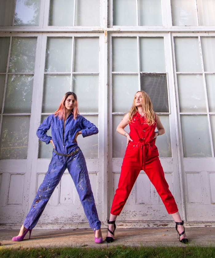 Two women posing confidently in vibrant jumpsuits against a vintage backdrop.