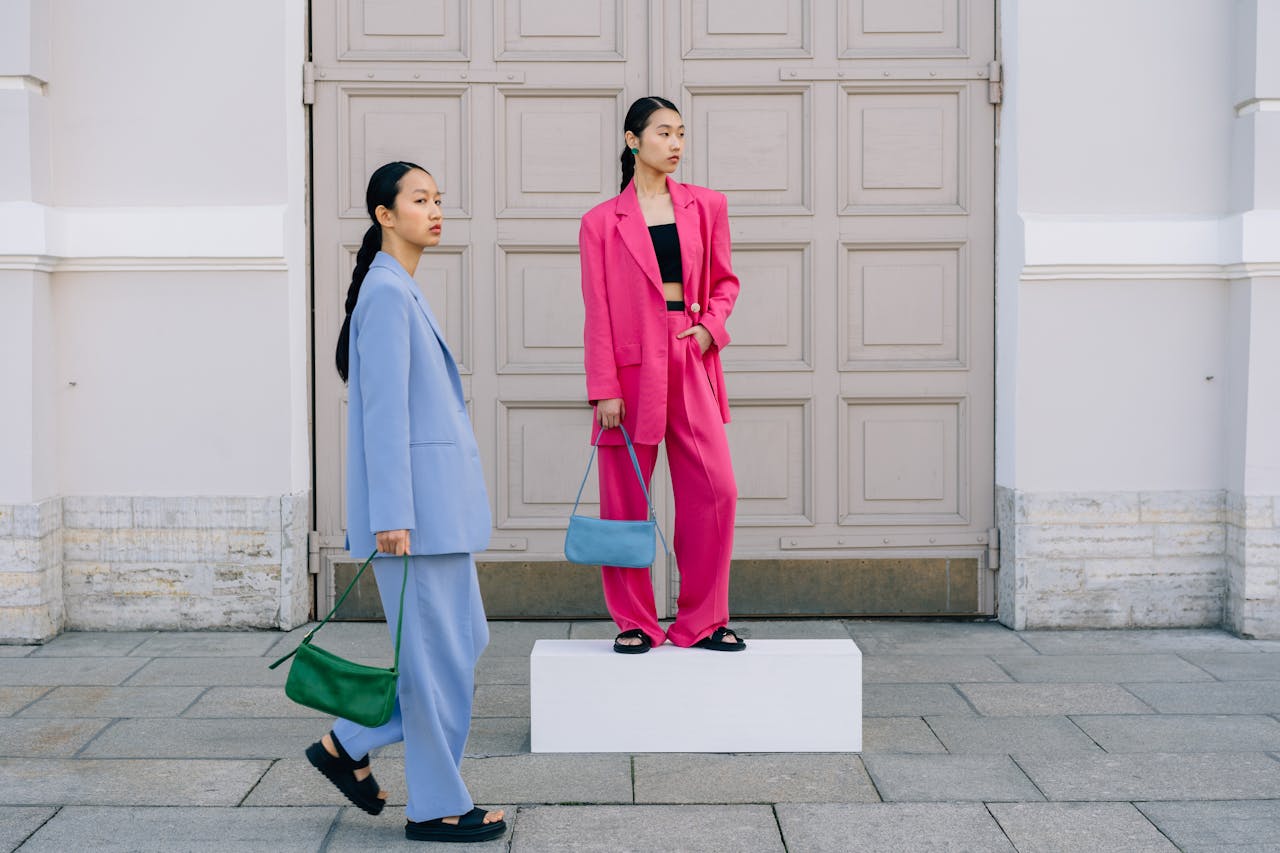 Two fashionable women in vibrant blazers pose with stylish handbags outside a vintage building.